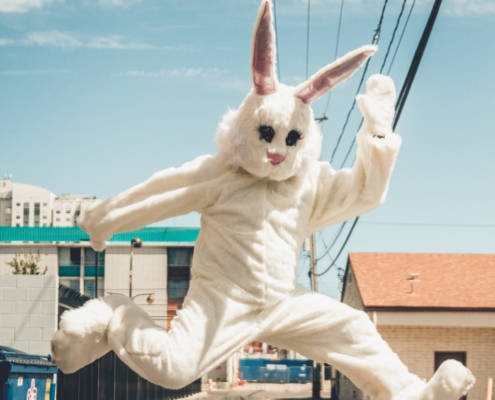 Person in a white bunny costume jumps energetically in an urban alley, with buildings and clear skies in the background.